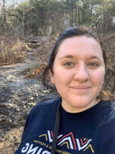 Adria takes a selfie while on an outdoor trail in autumn. She has her hair pulled back into a ponytail and is wearing a tshirt and crossbody bag. The plants along the trail are browned with some green leaves peeking through in the background.