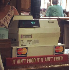 An old, white cash register at a restaurant. The total has come to $7.69 and the MasterCard logo is visible on the base of the register, most likely to show what cards are taken. Underneath the register is a wooden sign that has been painted red that says, "it ain't food if it ain't fried."