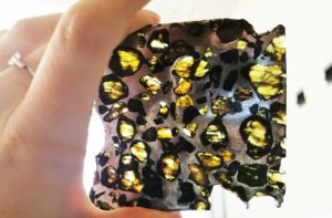 A white hand holding a meteorite slice up to a window. The meteorite is metallic with many gemstones; it is similar to a stained glass window. The gems are green/amber-colored.