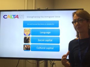 Author standing in front of digital display with text "Strengthening the Immigrant Voice: art and personal narratives as catalysts for language, social capital, and cultural capital.