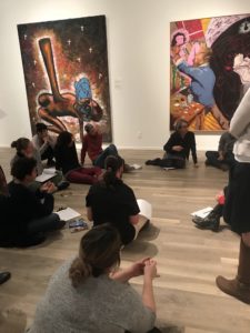 Eleven women sitting on the floor of an art museum gallery space having a discussion.