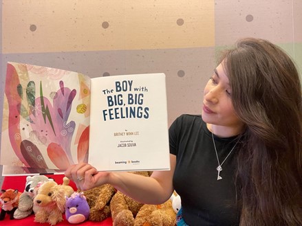 Asami Robledo-Allen Yamamoto, a light skinned Latina woman with long, wavy brown hair, is holding up an open, colorful book that reads 