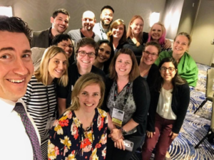 16 people gather in a cluster for a selfie photo at a conference.