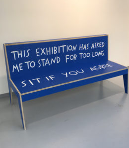 A bright blue bench. Hand-lettered text runs across the back and seat. It reads, “This exhibition has asked me to stand for too long. Sit if you agree.”