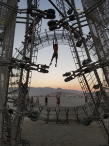 Image description: A man hangs by his arms from within a sculpture made of connected shopping carts that form a large circle approximately twenty feet high, and is installed in the desert.