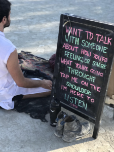 Image description: A man sits cross-legged in the desert next to a sandwich board that reads “Want to talk with someone about how you’re feeling , or share what you’re going through? Tap me on the shoulder; I’m here to listen.”