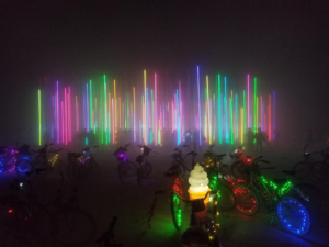 Image description: Tall neon lights of various colors and sizes rise vertically from the ground in the background, with bicycle adorned with different lights are in the foreground. The picture is taken at night in the desert.