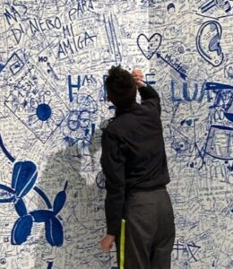 A person is shown writing on a wall with a blue pen. The walls and the floor have already been written on numerous times and he is adding to it. Although much of the writing is very small and unreadable, one can see large drawings, such as a Koons dog statue and a pierced ear.