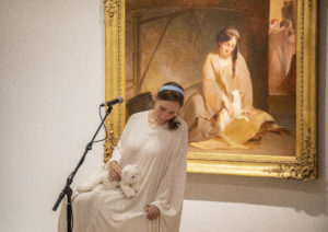 An actor with long, dark hair, pulled back by a headband, sits on a stool in front of a gold-framed painting. The actor wears a white gown and is holding a stuffed cat. The actor’s outfit and pose mirrors what is depicted in the painting. There is a microphone on a stand in front of the actor.