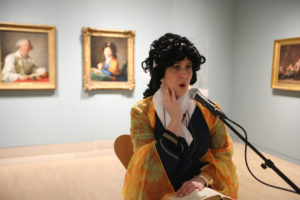 An actor with dark, curly hair and an orange wrap stands in a museum gallery in front of a gold-framed painting. The actor is speaking into a microphone.