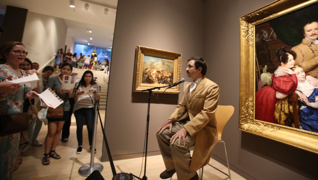 An actor sits in a museum gallery with a microphone in front of them. The actor has short brown hair, a moustache, and is wearing a tan suit and pants. There are people, who are holding pamphlets and pencils, gathered in front of the actor. To the actor’s right is a painting depicting what looks to be two women in dresses and a man wearing an outfit similar to that of the actor.
