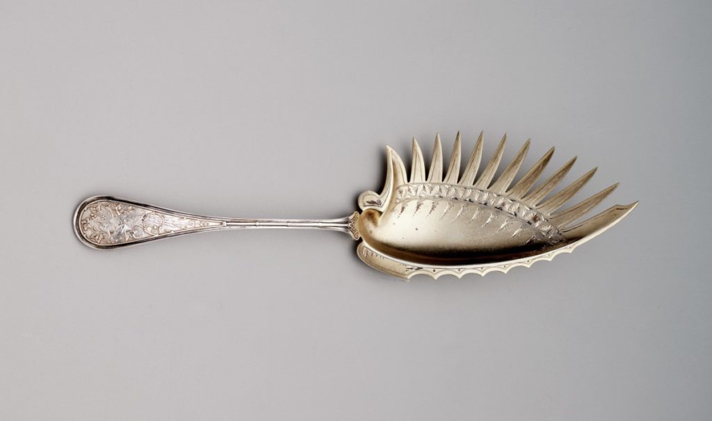 A silver utensil has a long stem with bird patterns on the bottom. The top serving portion has a concave oval that is surrounded on one side by long, sharp, feather-like points, while the other side has small half-circled dips.