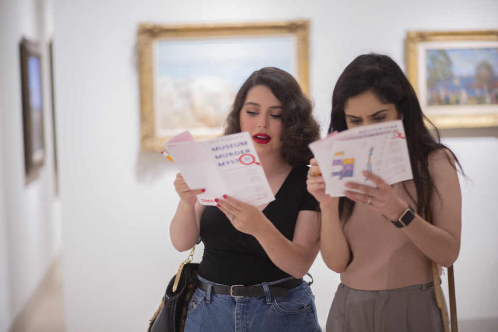 Two individuals stand in a museum gallery examining pamphlets labeled “Museum Murder Mystery.” They each have long dark, and one wears jeans and a black t-shirt, while the other wears grey pants and a dusty peach shirt.