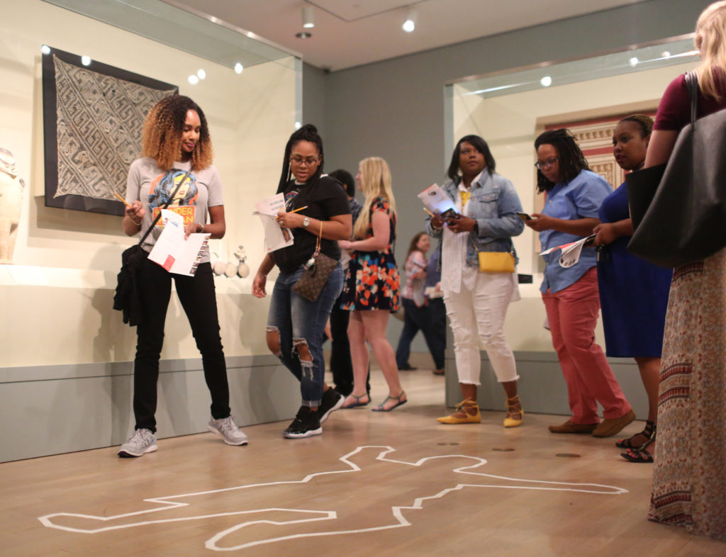 A group of people, holding pamphlets and pencils, stand in a gallery displaying textiles. They are gathered around a white traced outline of a body on a wooden floor. 