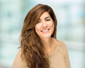 A woman with long brown hair is shown smiling. She wears a tan shirt. The background has been blurred.