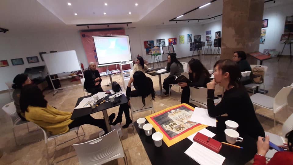 A group of individuals sit at tables grouped together. They are looking toward someone who is speaking. The classroom around them has art hung on the walls, while easels and white boards are scattered around.