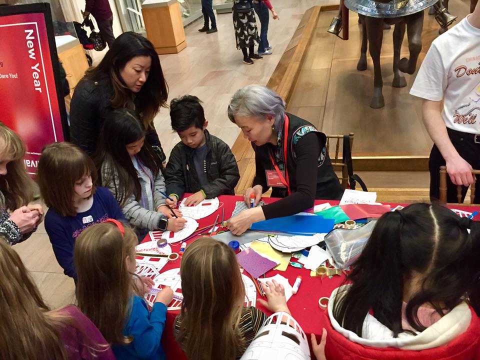 A group of children gather around a table. A person with grey hair is leading the children in an art activity. 