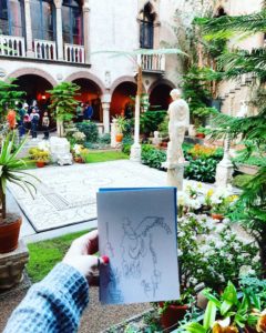 A white hand holding a line drawing of a sculpture in a courtyard. The courtyard is filled with many plants and shrubs, several classical marble sculptures, and shows a balcony with windows in the background.