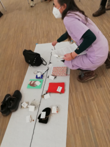 A woman, holding a pencil and a book, is crouching on the floor next to a long white sheet of paper on which objects are arranged along a middle axis formed by a black cord. The objects include things like shoes, bags, notebooks and a package of paper tissues. Small labels with handwritten notes are tied to most of the objects.