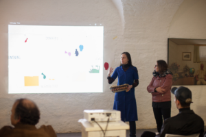 Two women stand next to a big sized white paper mounted on a white brick wall and illuminated by a beamer. On the paper multi-coloured cut out figures with different abstract and figurative shapes are arranged in a coordinate system spanning between the German terms Jenseits, Normalität, Einfühlung, and Gewalt, which translate to Beyond, Normality, Empathy, and Violence. One of the women is talking to an audience of which the heads of two people sitting in the first row are seen from the back.