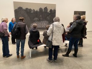 In this image, 11 people are looking at a very large, approximately 12-foot x15-foot painting. Some are standing; some are seated on a white plastic bench. All have their backs to the camera, except for the group leader who is looking at the group. Twelve dark gray ghost-like images in the painting are looking at one figure lying on a bed, except for one small child whose back is toward the bed. The figure in the bed is covered with a quilt that has some color in it, which is the only color in the painting. The viewer can’t tell if the figure in the bed is dead or alive.
