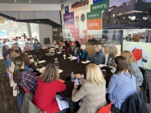 In this image, 21 people are seated around a long wide table, with water bottles and coffee cups in front of them. A few have remnants of a lunch. One person, in the back left corner is speaking; the others are all turned facing the speaker. The wall behind the table has posters of the museum exterior and inspirational banners. Some words on the banners are behind visitors at the table; however, the visible words read: “If it opened eyes, sparked conversation, stoked pride,” and “…memory, created a smile.”