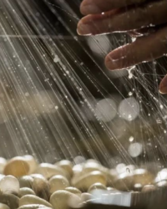 Water falls in a light sprinkle over lightly colored circular pebbles.On the upper right hand side, a hand reaches forward to touch the water before it can fall on the rocks.