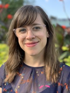 A photo portrait of a white woman with shoulder length hair and glasses with a blurred outdoor garden setting in the background.