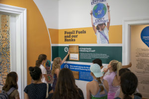 A group of ten college age students gather around an exhibition, whose wall text reads "Fossil Fuels and our Banks." One young woman with long blonde hair and a plaid dress lifts up an interactive text panel to reveal an answer, although the question and answer cannot be read clearly in the picture.