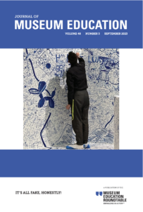 The cover of the Journal of Museum Education 48.3 featuring a person is shown writing on a wall with a blue pen. The walls and the floor have already been written on numerous times and he is adding to it. Although much of the writing is very small and unreadable, one can see large drawings, such as a Koons dog statue and a pierced ear.