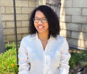 Aubrey is seated wearing a white button down shirt, glasses, with a smile and shoulder length brown curly hair.