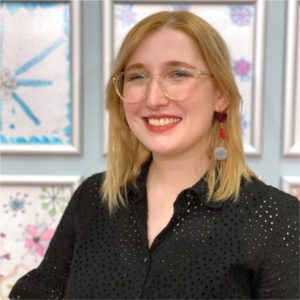 Hazel Theriault's headshot. A blonde woman with shoulder length hair and glasses smiles in front of some art work.