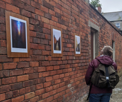 A person with blonde hair looks to the left at a red brick wall, upon which are hung several pieces of art.