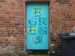 An image of a turquoise door set into a red brick wall dominates this image. The door has large letters spelling "EGRESS" that are entwined with illustrations of flowers. and leaves.