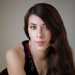 A woman with long black hair looks at the camera. Behind her is a blank wall.