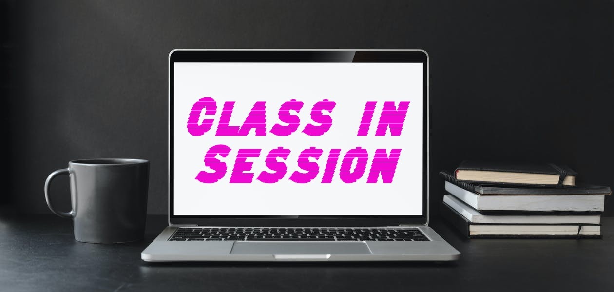 A silver laptop sits on a table with a grey mug to the left of the screen and notebooks and books to the right. The screen reads “Class in Session” in pink letters.