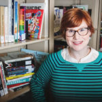 Headshot of Kristin. She is a white woman with short red hair with glasses. She is smiling at the camera with several bookshelves full of books behind her. 