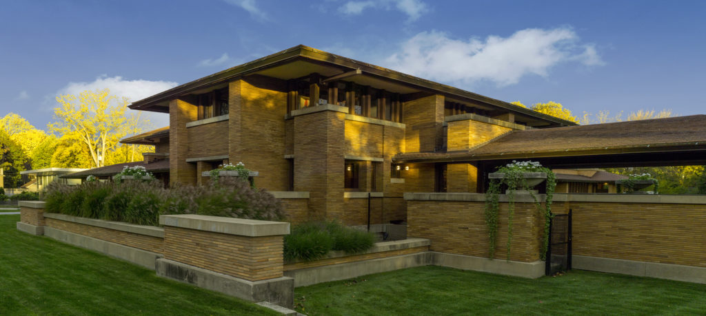 Photograph taken at sunset of Frank Lloyd Wright’s Martin House, showing the two story building’s Prairie Style of architecture with long, low roofline and facade of stone and wood in natural colors. A well maintained yard with planters and flowers and freshly cut grass line the front of the building.