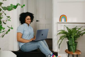 A woman in jeans and a blue top sits on an indoor step watching a laptop. She is surrounded by indoor plants and a rainbow artwork