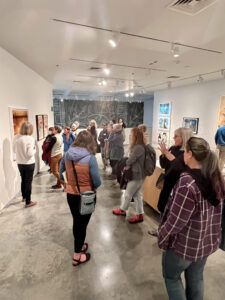 In this image, about 15 book club members in a museum gallery that’s about 25-feet wide by 40-feet long are facing a painting on the wall. The gallery walls and ceiling are white. The docent leader has her back to the camera and is looking at the painting. All you can see of the painting is reddish-yellow at the top and bottom with a dark black band through the middle. There are several other paintings on the walls, but they are too far from the camera to be recognizable.