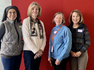 This image has 4 women, two on the left, two on the right, facing each other at a slight angle, and turned toward the camera. The women are wearing vests, sweaters and scarves.